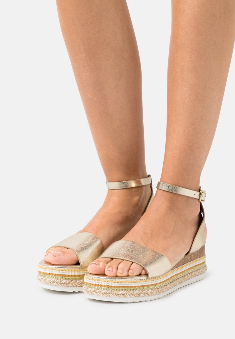 Pieds portant des sandales compensées dorées métalliques avec des brides à la cheville et des détails en corde tressée sur les semelles compensées sur fond blanc.