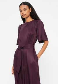 Vestido de cetim roxo com mangas curtas, cintura marcada com um laço de cinto e bolsos laterais; textura suave e caimento elegante.