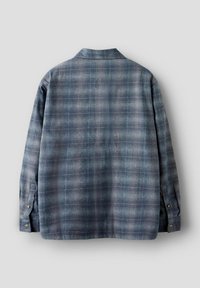 Sinine ruuduline särk, mis on valmistatud pehmest kootud kangast tekstuurse viimistlusega, iseloomustab lõdvestunud lõiget, pikki varrukaid, kraega ning nööpidega mansette.