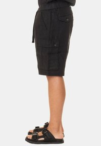 Pantaloni cargo neri in lino, lunghezza al ginocchio, con tasche laterali, vita con coulisse e dettagli in bottone. Indossati con sandali neri con cinturini.