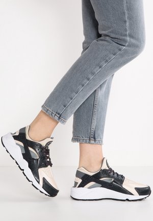 Nogi w jasnoniebieskich dżinsach i czarnych, beżowych oraz białych sportowych butach, jedna stopa lekko uniesiona na białym tle.