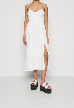 Frau trägt ein weißes Midikleid mit dünnen Trägern und seitlichem Schlitz, kombiniert mit weißen Plateau-Sandalen mit doppelten Schnallen.