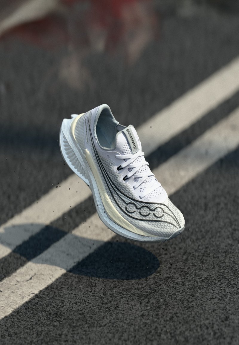 Λευκό αθλητικό παπούτσι Saucony με μαύρο λογότυπο, αιωρούμενο πάνω από ασφάλτινο δρόμο με λευκή γραμμή.