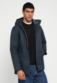 Sötétkék, vízálló kapucnis dzseki kockás béléssel, elöl cipzárral és oldalzsebekkel. Fekete, kötött mintás pulóverrel párosítva.