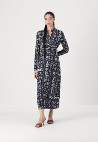 Lungo abito blu navy con motivi floreali e geometrici bianchi, colletto, maniche lunghe e vita stretta. Abbinato a sandali con tacco marroni.