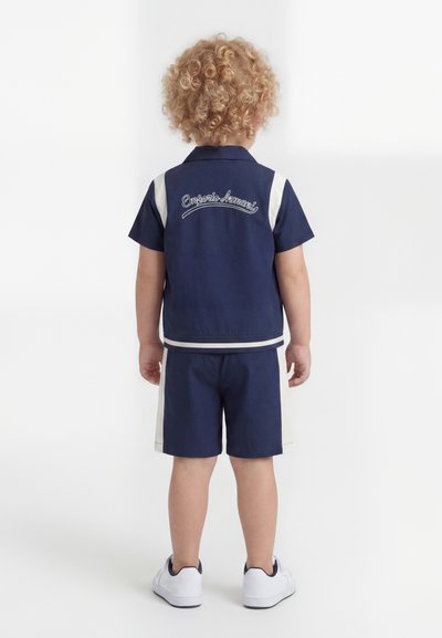 Παιδί με σγουρά ξανθά μαλλιά που φοράει μπλε-σκούρο και λευκό πουκάμισο και σορτς Emporio Armani, στέκεται με την πλάτη γυρισμένη σε λευκό φόντο.