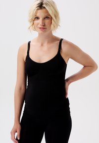 Blonde zwangere vrouw met kort haar, gekleed in een zwart mouwloos zwangerschapshemd, met haar hand op haar rug rustend, kijkend naar de camera.