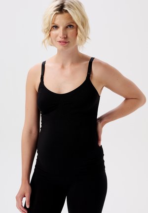 Blonde pregnant woman with short hair wearing a black sleeveless maternity top, hand resting on back, looking at camera.