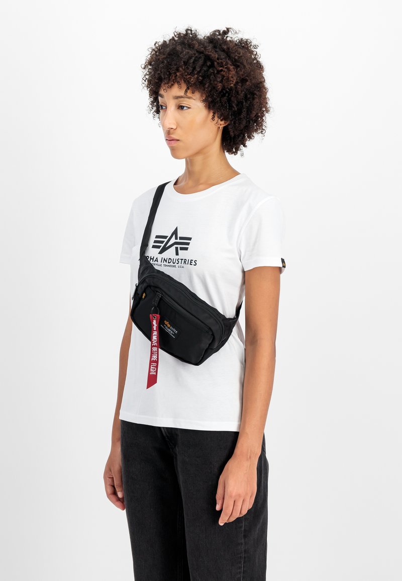 Black fabric crossbody bag with a zippered front pocket and a red strap featuring text. The model wears a white shirt and black jeans.