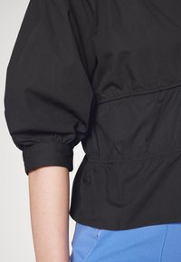 Svart cropped jacka med ett samlat midja och puffy ärmar. Slät textur och elastiska manchetter. Bärs över ljusblå byxor.