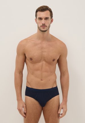 Spierachtige man die voor een effen achtergrond staat, draagt marineblauwe slip, kijkt recht vooruit met een neutrale uitdrukking.