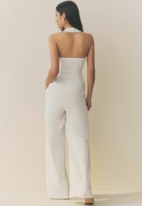Salopette blanche avec un design sans bretelles, dos smocké, jambes larges et corsage ajusté. Tissu lisse avec une texture unie.