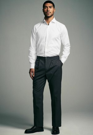 Camisa blanca de manga larga con cuello, combinada con pantalones oscuros de rayas finas y mocasines negros. La tela parece suave y estructurada.