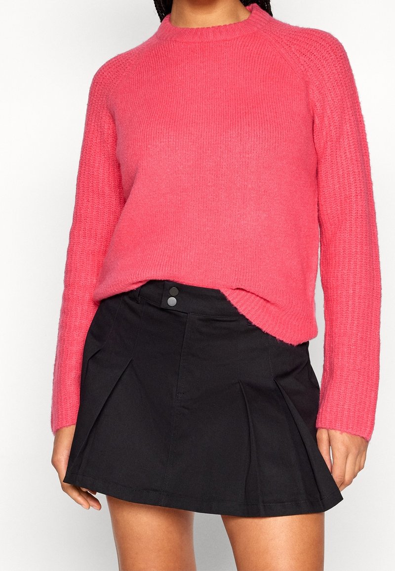 Femme portant un pull en maille rose vif rentré dans une mini-jupe plissée noire taille haute, debout devant un fond blanc.