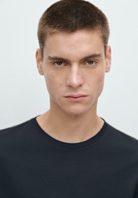 T-shirt nera a girocollo, tessuto liscio, design aderente, semplice e senza modelli, indossata da un modello con capelli corti e tratti del viso minimali.