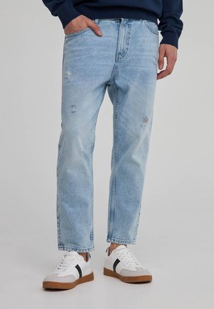 Mann trägt hellblaue, abgenutzte Jeans, dunkelblauen Pullover und weiße Turnschuhe mit schwarzen Streifen vor neutralem Hintergrund.