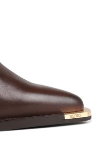 Punta de zapato de cuero marrón oscuro con suela plana y una punta metálica dorada grabada con la palabra "BRONX" sobre un fondo blanco.