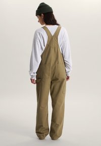 Olive green overall jumpsuit with loose fit, adjustable straps, and multiple pockets, paired with a gray long-sleeve shirt and green beanie.