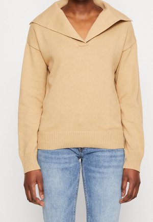 Beige strikket sweater med bred krave og ribstrikkede manchetter, båret med lyseblå jeans på en person vist fra nakke til hofter.