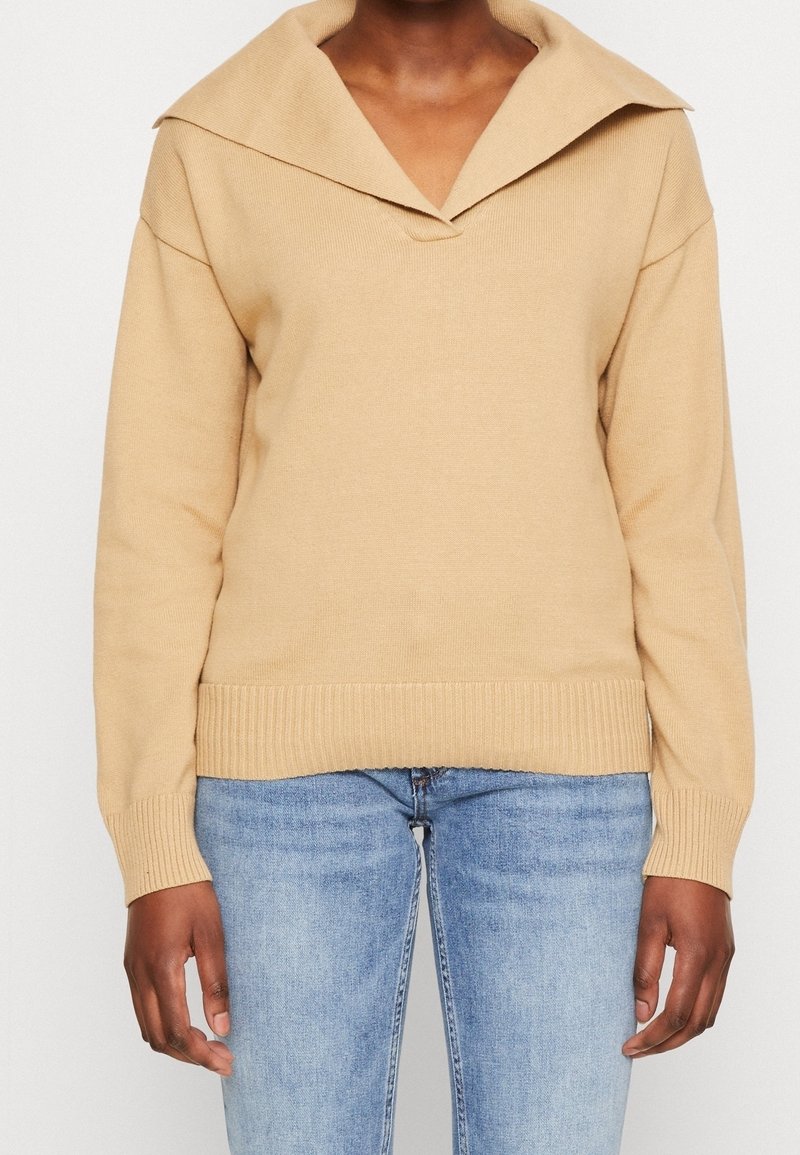 Pull beige en maille avec un large col et des poignets côtelés, porté avec un jean bleu clair sur une personne montrée du cou jusqu’aux hanches.