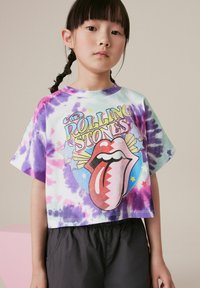 Kruopēta T-kreklu ar raibs zīmējums, kas ietver rozā, violetas un zaļas krāsas. Priekšā ir krāsains "Rolling Stones" logotips ar mēli.