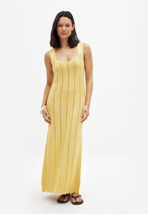 Gelbes, gestricktes Maxikleid mit quadratischem Ausschnitt, das ein Zickzackmuster und gerippte Akzente aufweist. Getragen mit braunen Flip-Flops.