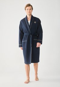 Marineblauer Bademantel mit Waffelstruktur, einem Schalkragen, goldfarbener Paspelierung und zwei vorderen Taschen. Mit einem gestickten Logo auf der Brust.
