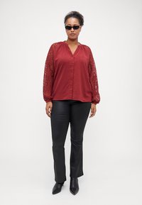 Bordová blůza s krajkovými rukávy, zapínání na knoflíky, v kombinaci s těsnými černými kalhotami a kotníkovými botami. Modelka má na sobě tmavé sluneční brýle.
