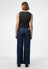Kvinna som står med ryggen mot kameran, iklädd en ärmlös svart topp, vida mörkblå jeans och svarta skor mot en neutral bakgrund.