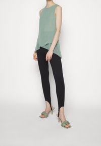 Helles grünes ärmelloses Top mit schichtigem Design, kombiniert mit schwarzen Leggings mit einzigartigem Saum und grünen gewebten Absatzsandalen.