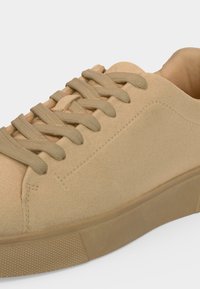 Baskets basses en daim beige avec lacets assortis, col rembourré et semelle en caoutchouc texturée sur fond clair.