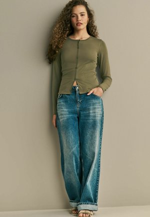 Olivgrünes Langarmoberteil mit Knopfverschluss und Rundhalsausschnitt, kombiniert mit lockeren blauen Denim-Jeans mit einer ausgewaschenen Optik und weitem Bein.