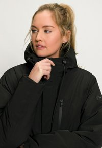 Veste noire isolante avec un col haut et une capuche ajustable. Comprend une poche zippée et une texture lisse. Gros plan sur la zone de la poitrine.