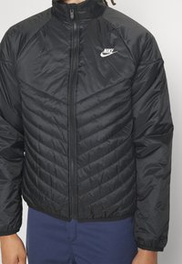 Černá péřová bunda s vysokým límcem, zapínáním na zip a vzorem v šipek. Obsahuje malé bílé logo Nike na levé straně hrudníku.