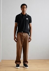 Zwarte polo met witte accenten, bruine wijde broek en zwarte schoenen met witte zolen. Eenvoudig, casual en een getailleerd ontwerp.