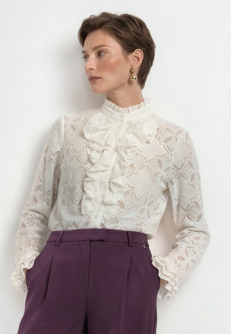 Woman with short hair wearing white lace ruffled blouse and dark purple high-waist pants, looking sideways against a plain light background.
