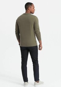 Olijfgroene shirt met lange mouwen, zachte textuur, ronde hals, getailleerd ontwerp. Draag het met een donkere spijkerbroek en witte sneakers, gezien vanaf de achterkant.