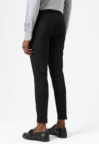 Zwarte op maat gemaakte broek met een smalle pasvorm, voorzien van een gladde textuur en omgeslagen pijpen. Gecombineerd met zwarte loafers en een grijs shirt met lange mouwen.