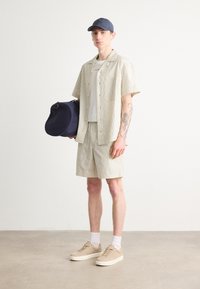 Camisa de manga corta a rayas y pantalones cortos a juego en beige claro, acompañados de un bolso de deporte azul marino y zapatillas beige con calcetines blancos.