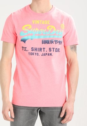 Man draagt een roze SuperDry vintage t-shirt met blauwe, gele en zwarte tekst, gecombineerd met grijze jeans, armen ontspannen naast het lichaam.