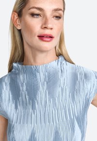 Top plissettato blu chiaro con collo alto e maniche corte, caratterizzato da un tessuto testurizzato e lucido e da pieghe verticali intricate.