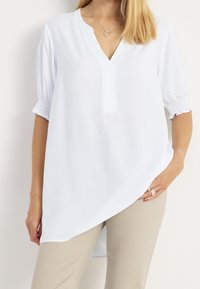 Blusa bianca leggera con scollatura a V, maniche corte arricciate e orlo asimmetrico. Abbinata a pantaloni beige, che mettono in risalto la camicia.