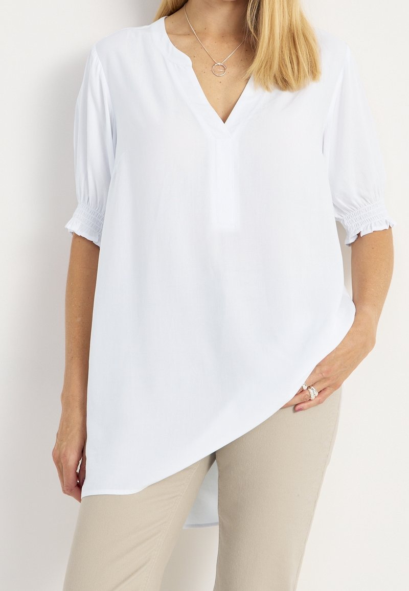 Blusa bianca leggera con scollatura a V, maniche corte arricciate e orlo asimmetrico. Abbinata a pantaloni beige, che mettono in risalto la camicia.