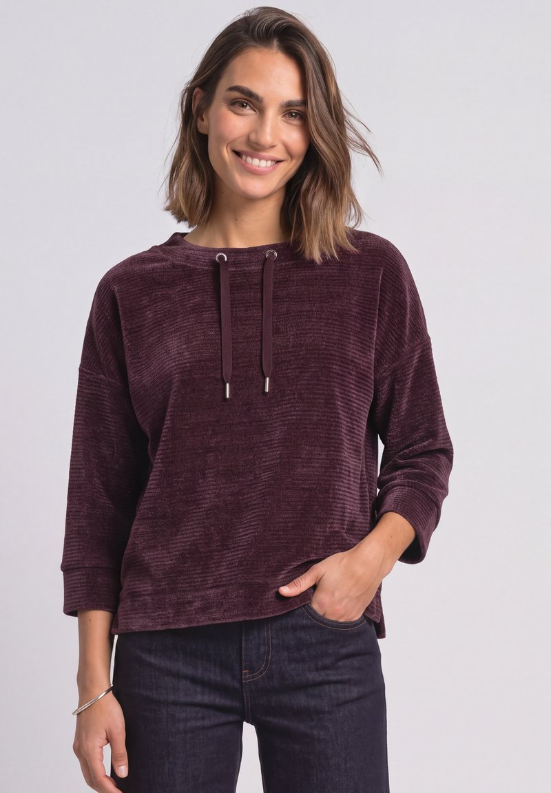 Femme souriante aux cheveux mi-longs, portant un haut à manches longues texturé violet foncé et un jean foncé, main dans la poche, sur fond clair.