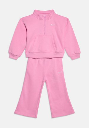 Różowy dres Nike dla małych dzieci z bluzą na długi rękaw z półzamkiem i szerokimi spodniami, z małymi białymi logo Nike swoosh na bluzie i spodniach.