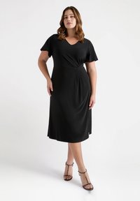 Zwarte jurk tot de knie met een V-hals, korte mouwen en een draai-detail bij de taille. Glad, lichtgewicht materiaal. Gepaarde met sandalen met bandjes en luipaardprint hakken.