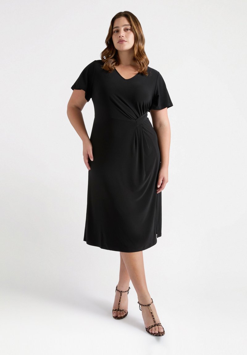 Zwarte jurk tot de knie met een V-hals, korte mouwen en een draai-detail bij de taille. Glad, lichtgewicht materiaal. Gepaarde met sandalen met bandjes en luipaardprint hakken.