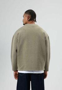 Lichtgroene oversized sweatshirt met een ronde hals, verlaagde schouders en geribbelde manchetten. Draagt over een wit shirt, gecombineerd met donkere jeans.