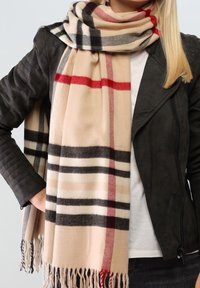 Femme portant une écharpe à carreaux beige avec des franges sur une veste en cuir noire et une chemise blanche, main sur la hanche devant un fond uni.