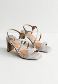 Sandales argentées à talons avec des brides croisées, bout carré et finition texturée. Elles disposent d'un talon chunky et d'une bride de cheville ajustable.